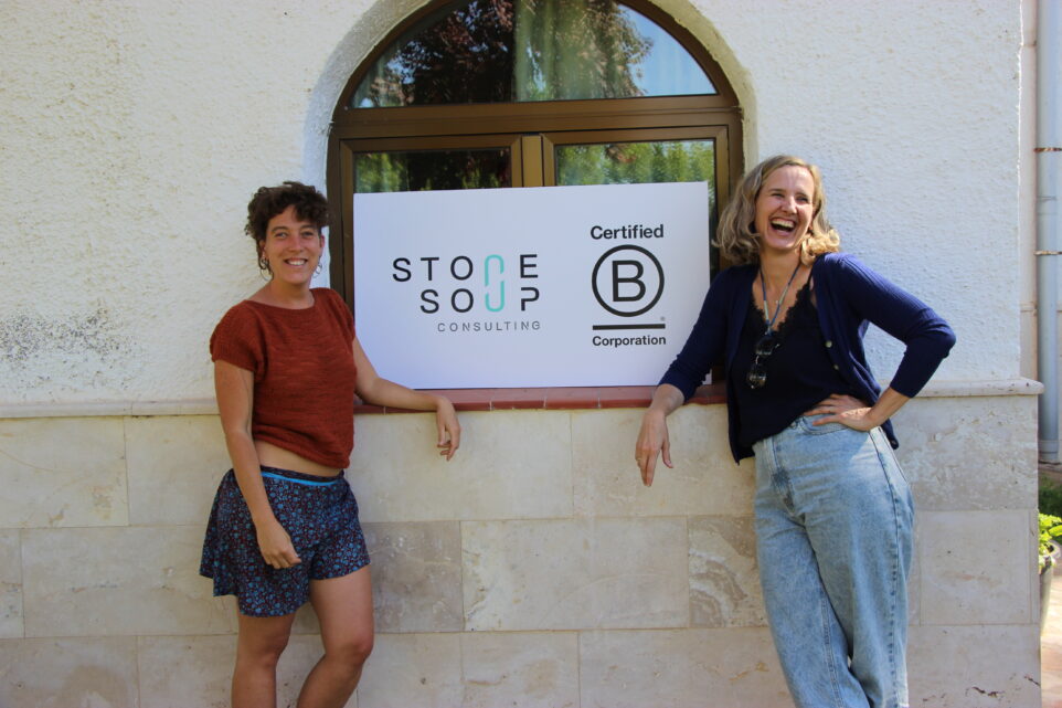 Two community female members stand next to a board with bcorp and stone soup logos