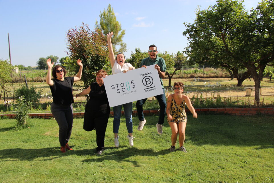 Five community members hold a board with stone soup and bcorp logo while jumping