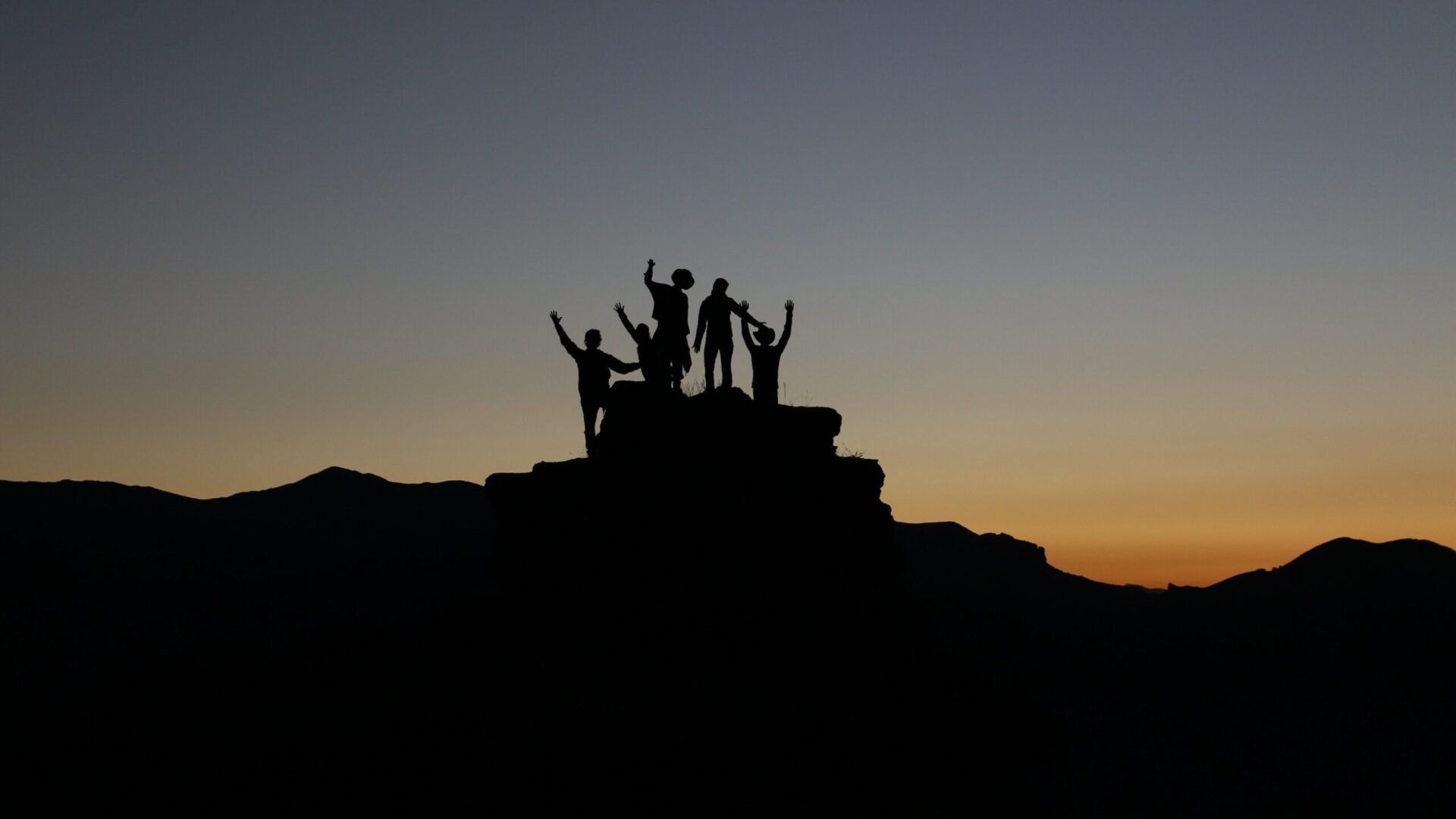 Group of people on top a rock with their arms extended, as in victory. It's the sunset, can only see shapes.